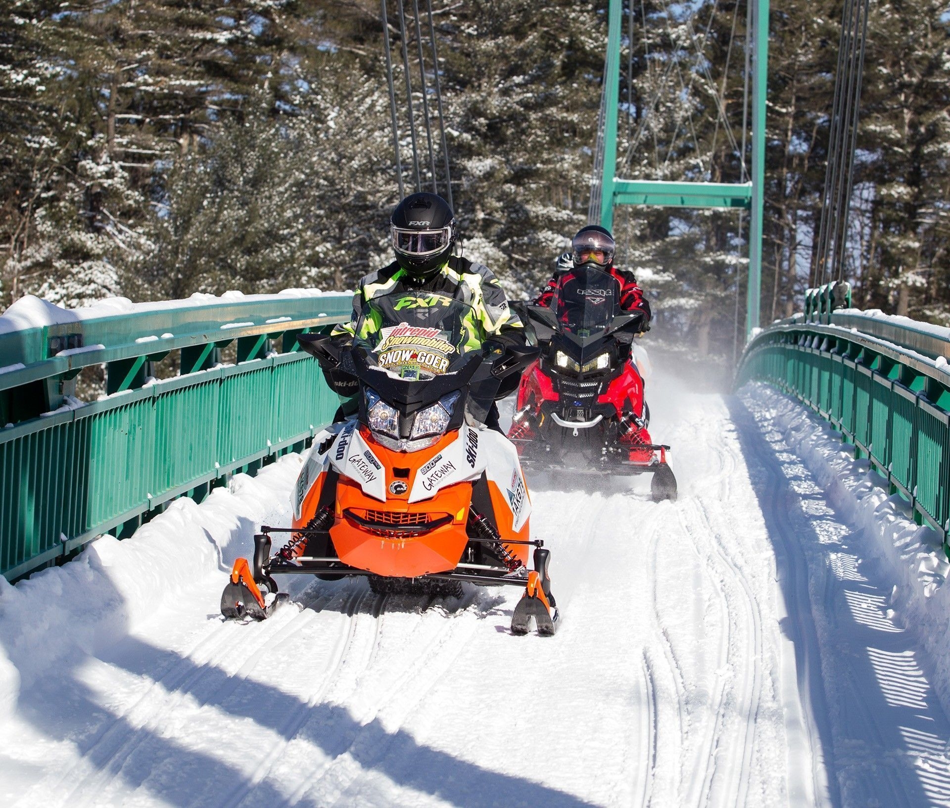 Two people on snowmobiles cross the green snowmobile bridge over the french river.