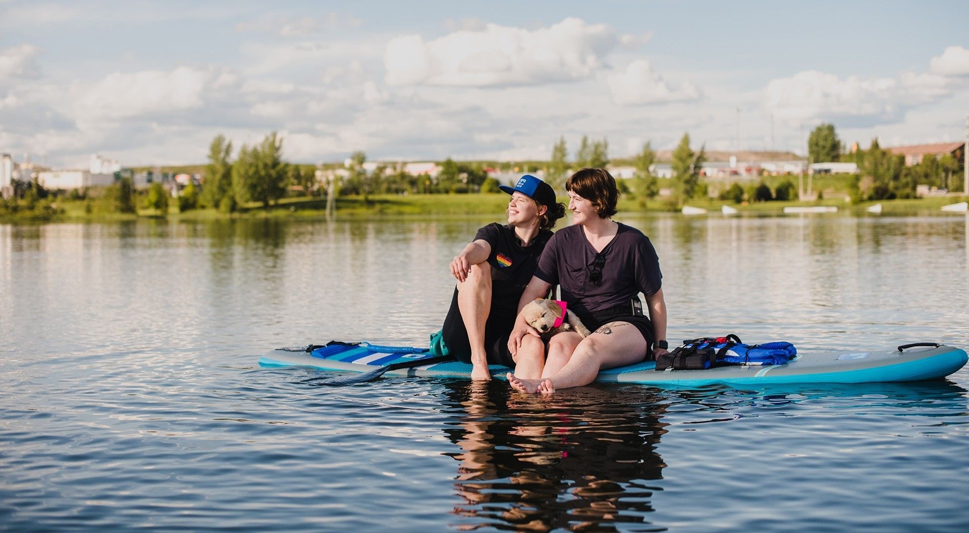 Two women sit side by side on a paddle board with a puppy on their laps.