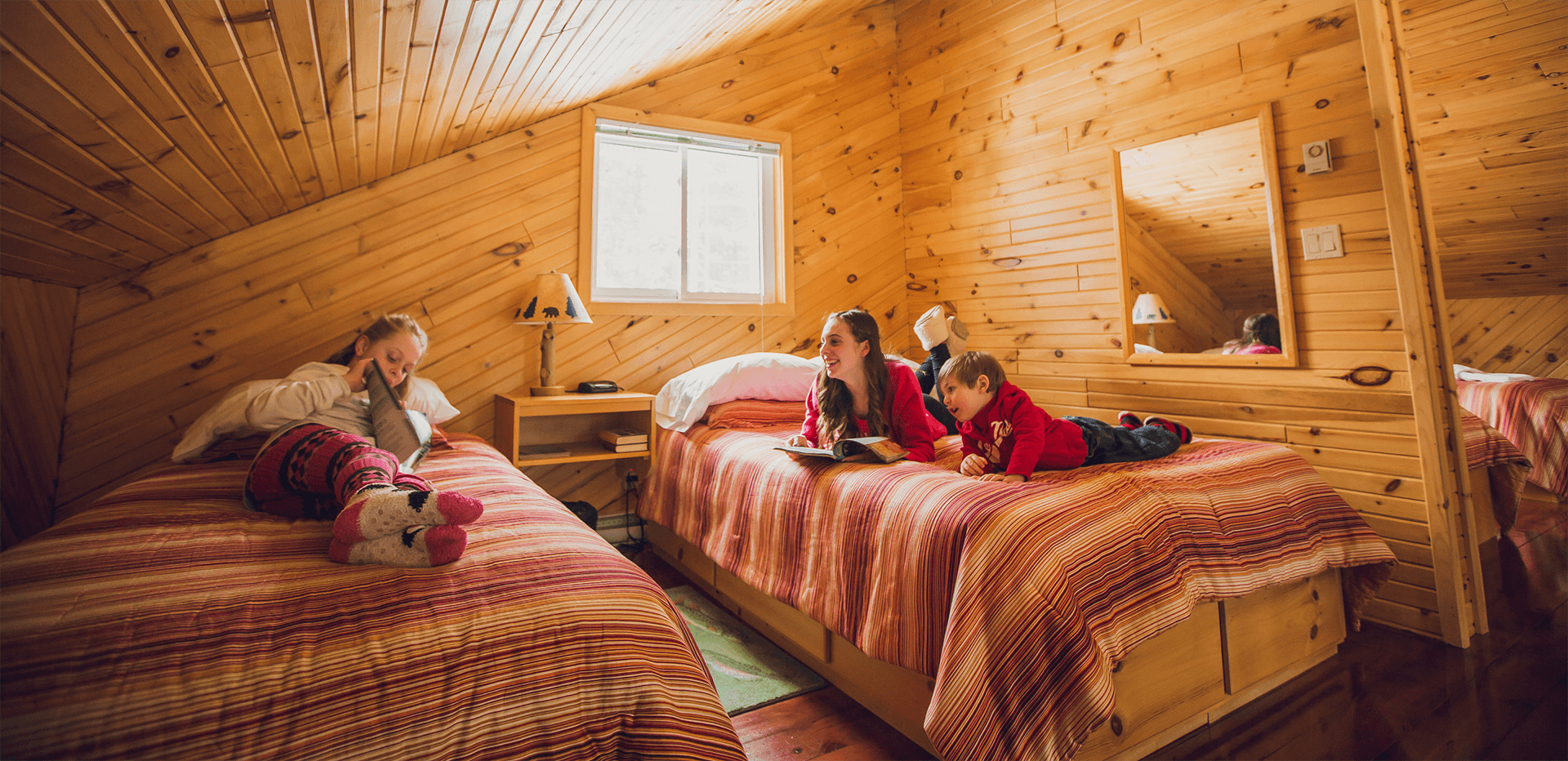 Three kids sit on comfy beds in a sunlit wooden cabin reading books and laughing.