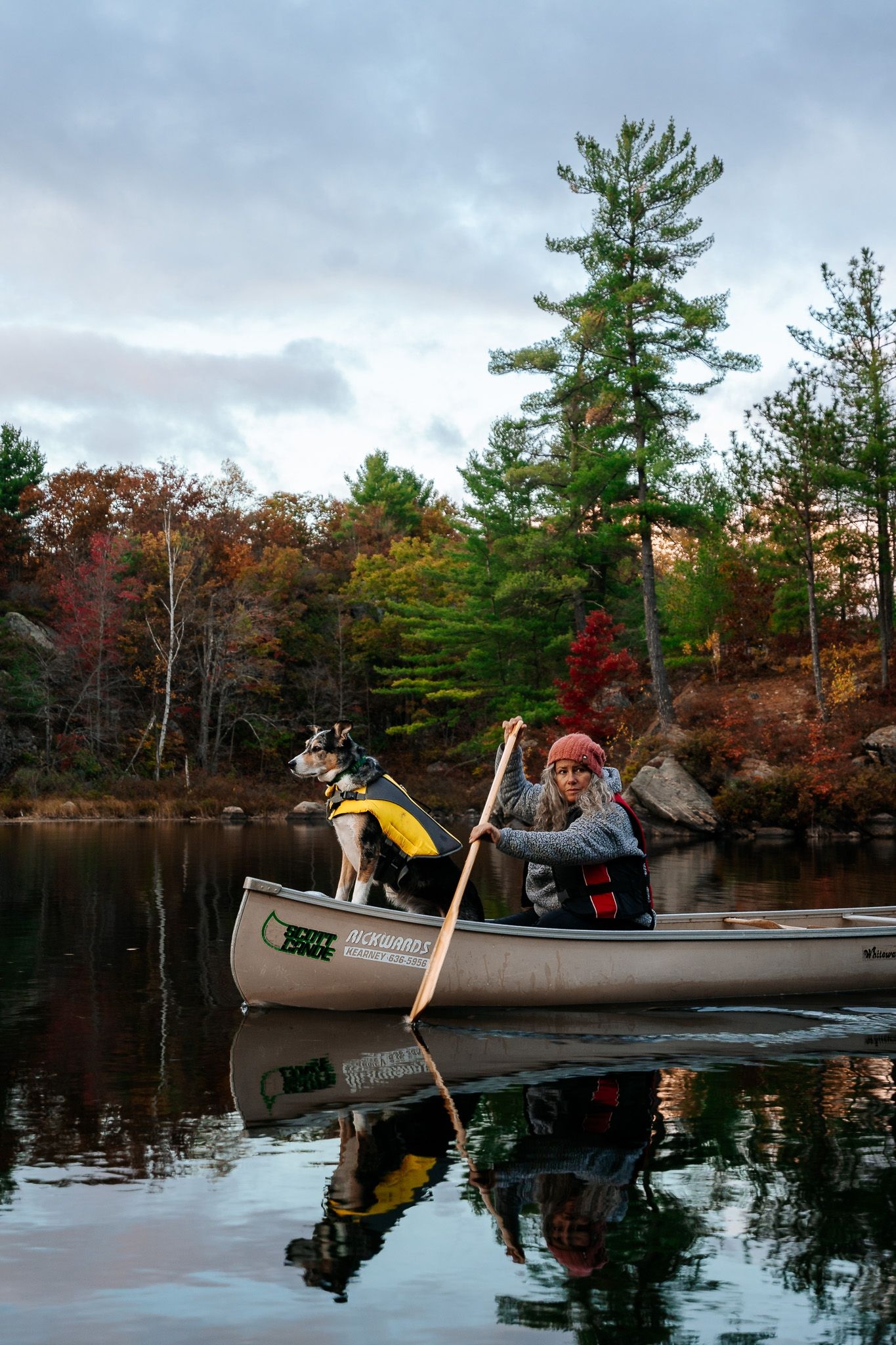 a woman paddles a canoe in the fall with a dog wearing a lifejacket in the bow