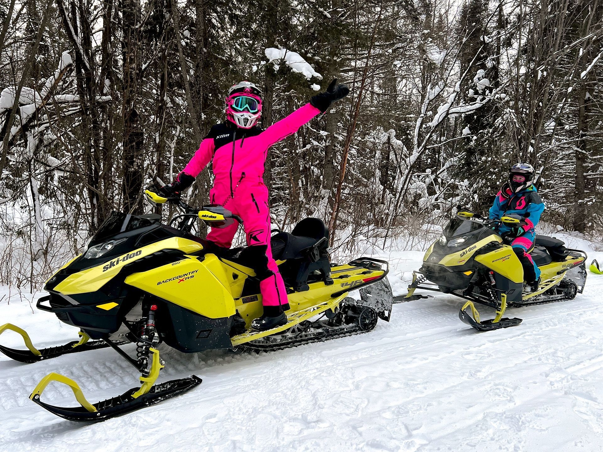 A two young girls dressed in pink and blue snow suits ride yellow snowmobiles on a snowy trail.