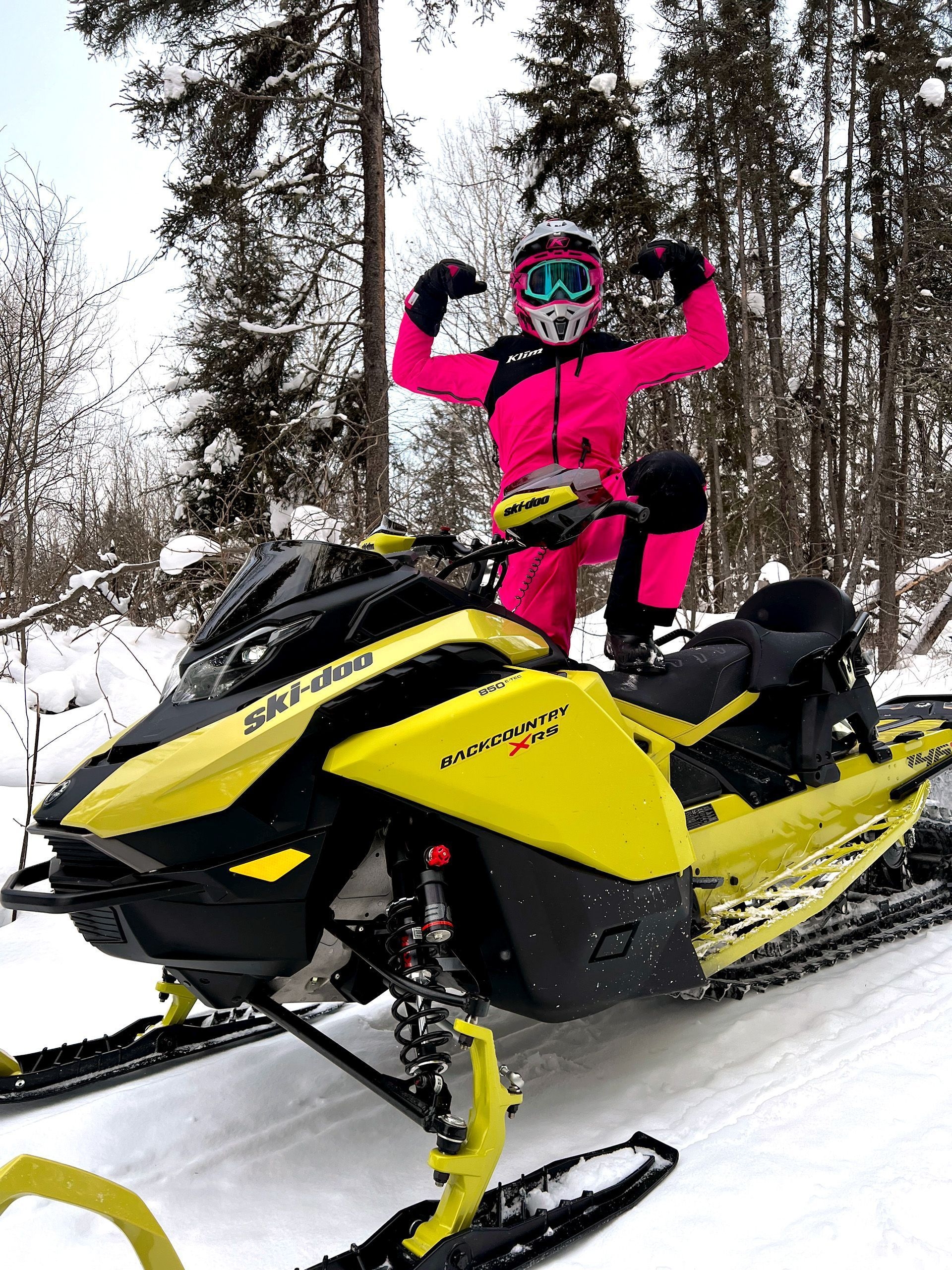 A two young girls dressed in pink and blue snow suits ride yellow snowmobiles on a snowy trail.