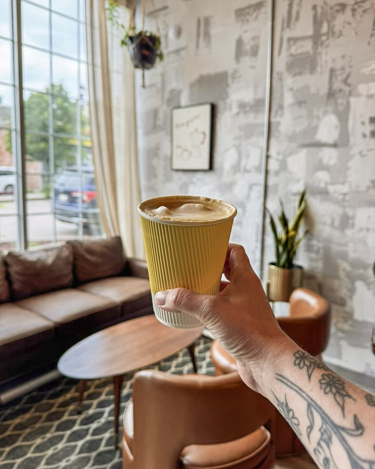 A tattooed arm holds out a latte in a yellow paper cup in front of a vintage couch sitting in front of a large picture window