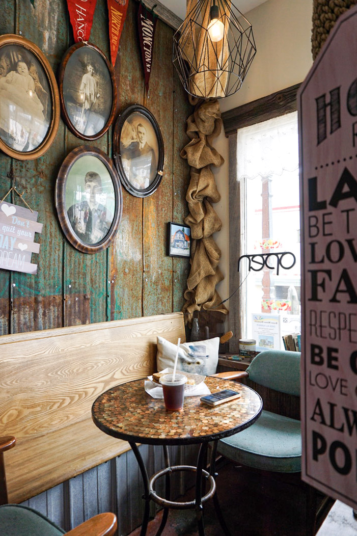 View of a cozy coffee shop with antique art on the wall and a coffee on a small wooden table in front of a bright window