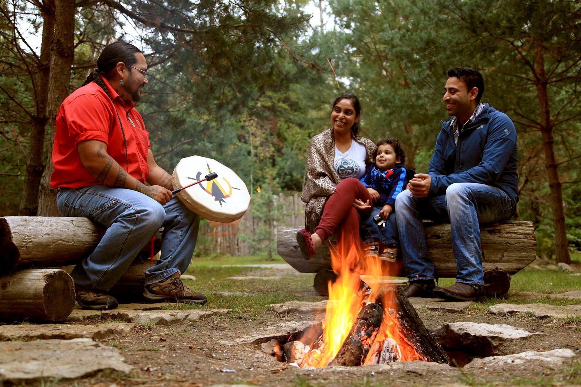 People sitting around a campfire looking at a man with a traditional drum.