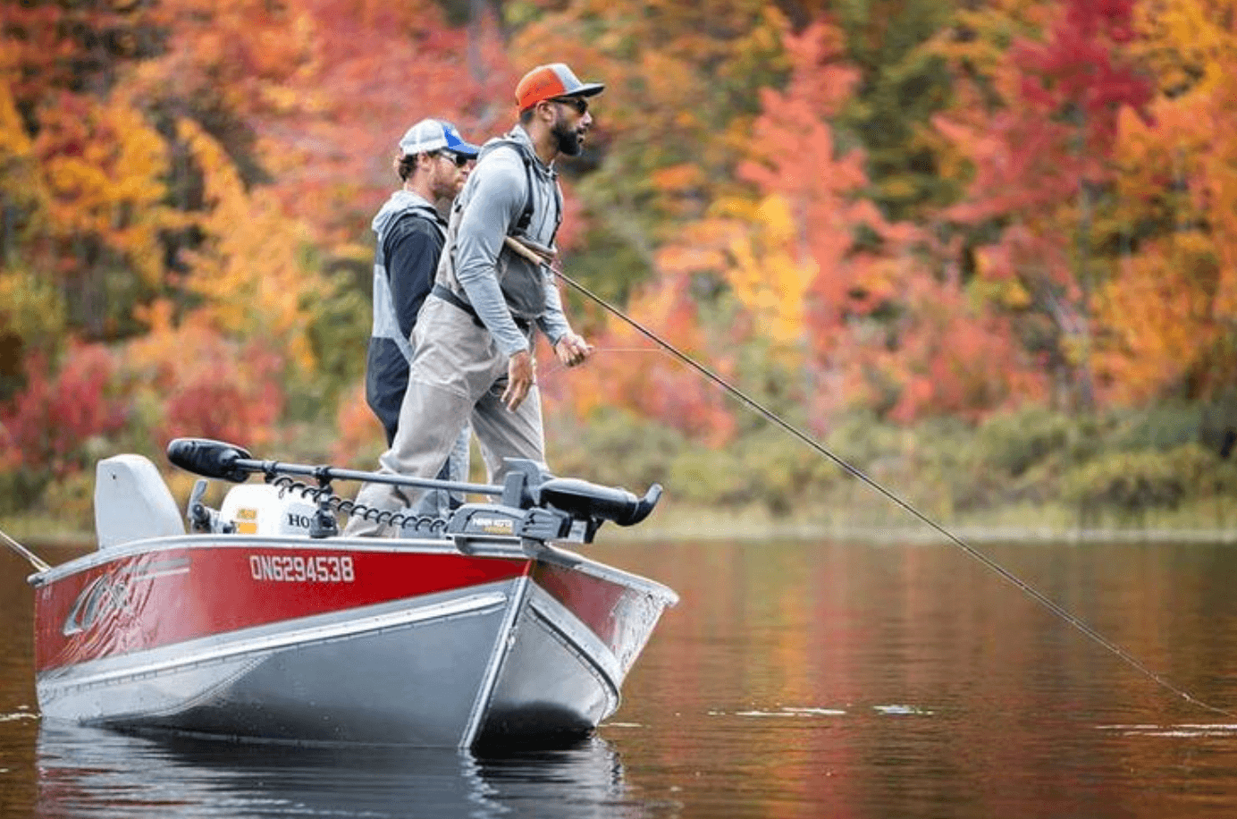 Two people fish out of a small fishing boat in front of fall colours.