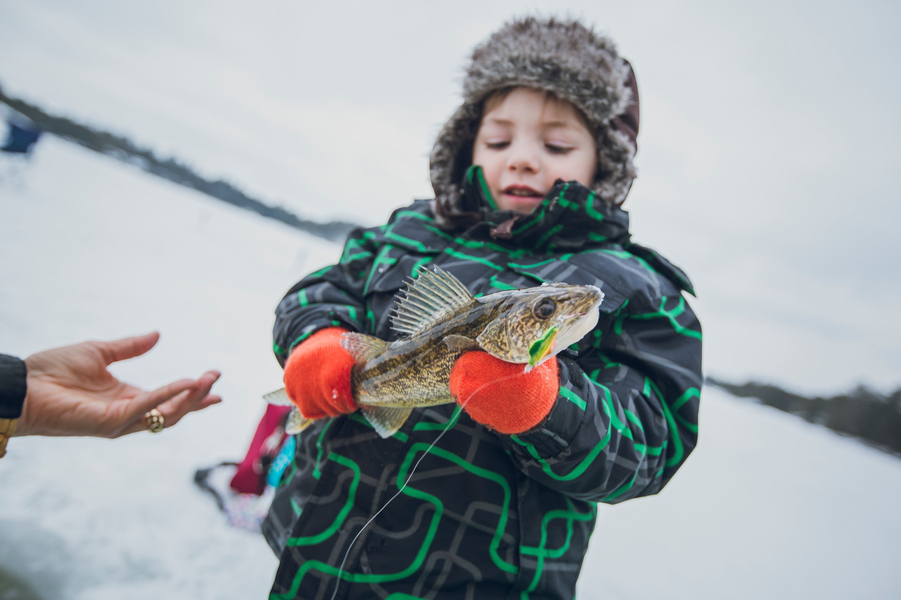 A kid in a green jacket and red mits holds a freshly caught fish.