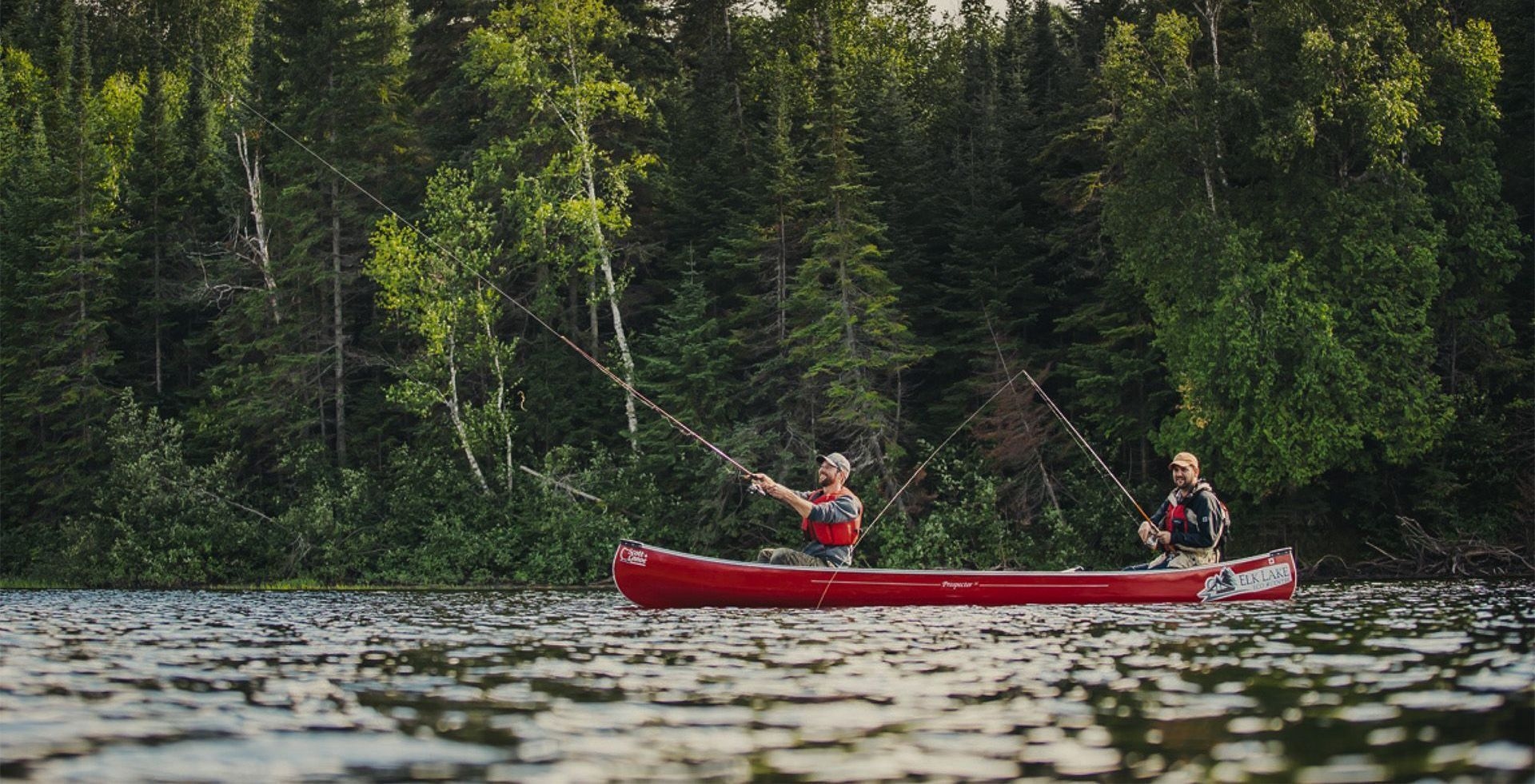 Two young people in fishing in a red canoe on a sparkling river, with tall conifers in the background.