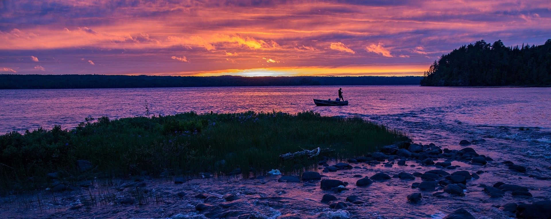 A person stands at the back of their boat casting a line in front of a pink, orange, and purple sunset.