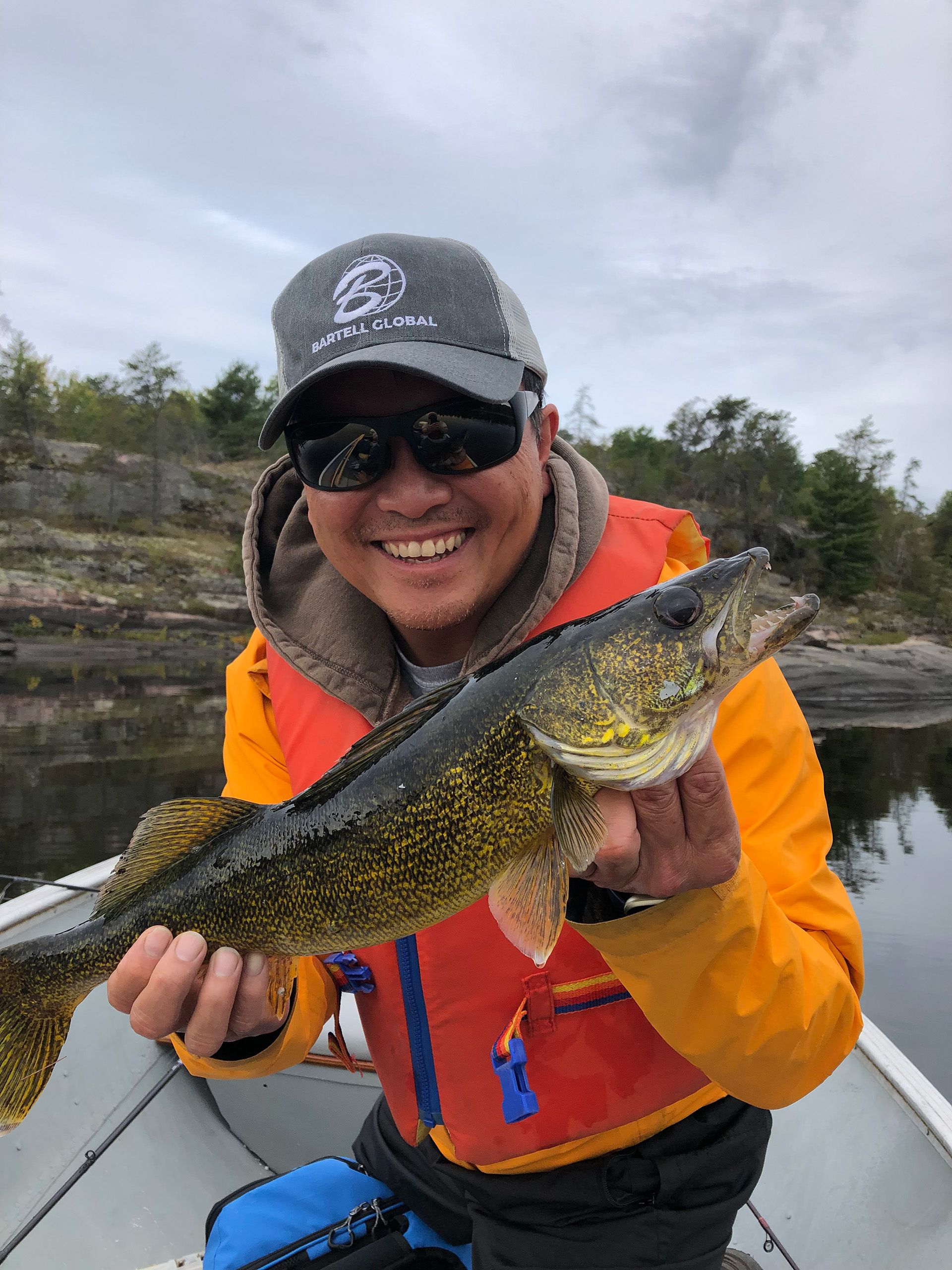 A person wearing a bright orange sweater and red lifejacket smiles and holds a medium sized fish