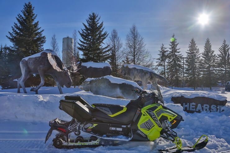 A green snowmobile is parked in front of the Hearst town sign, which also has a statue of a moose.