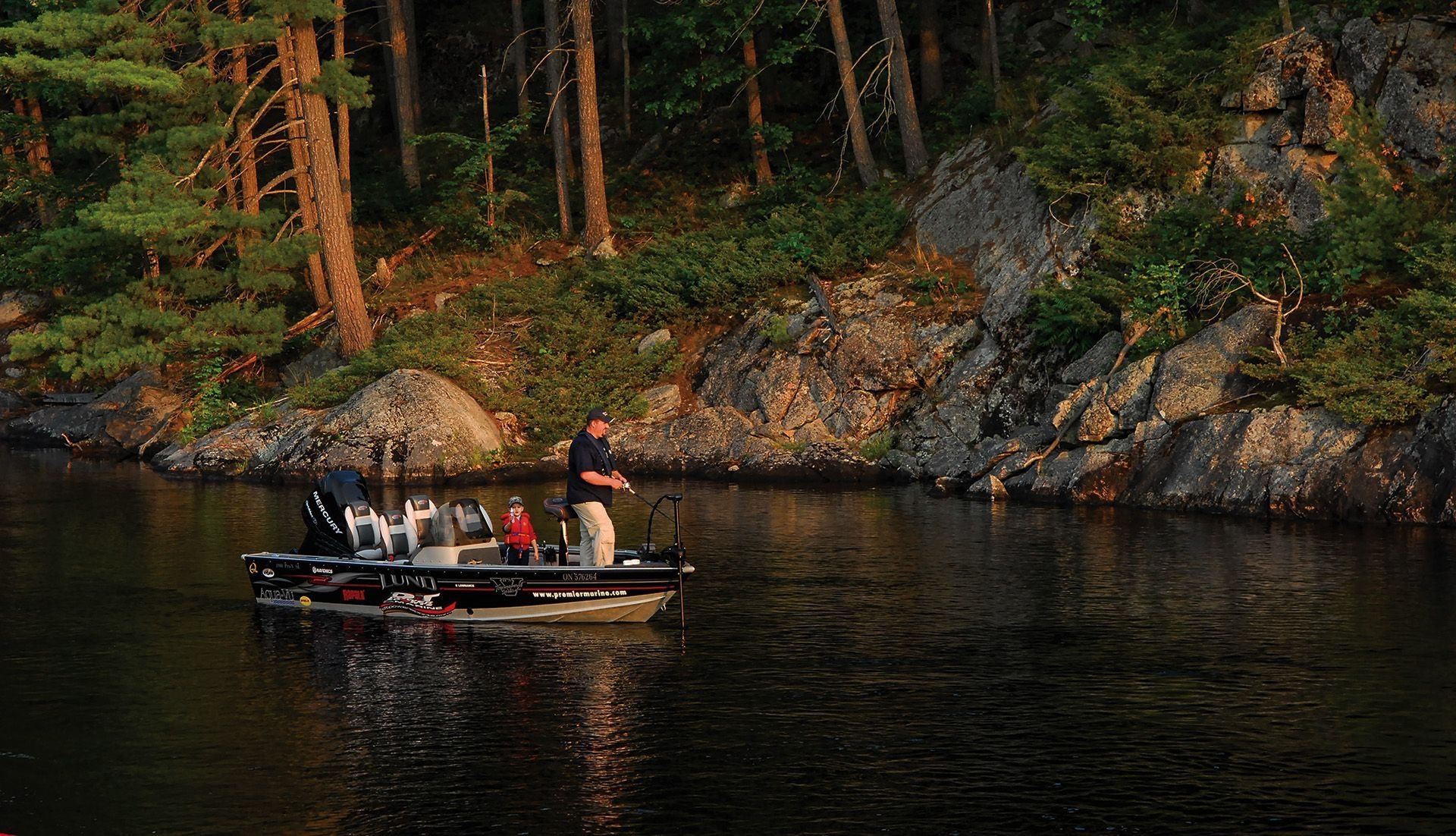 A man and his son are fishing out of a small motorboat close to a rocky and forested shoreline.