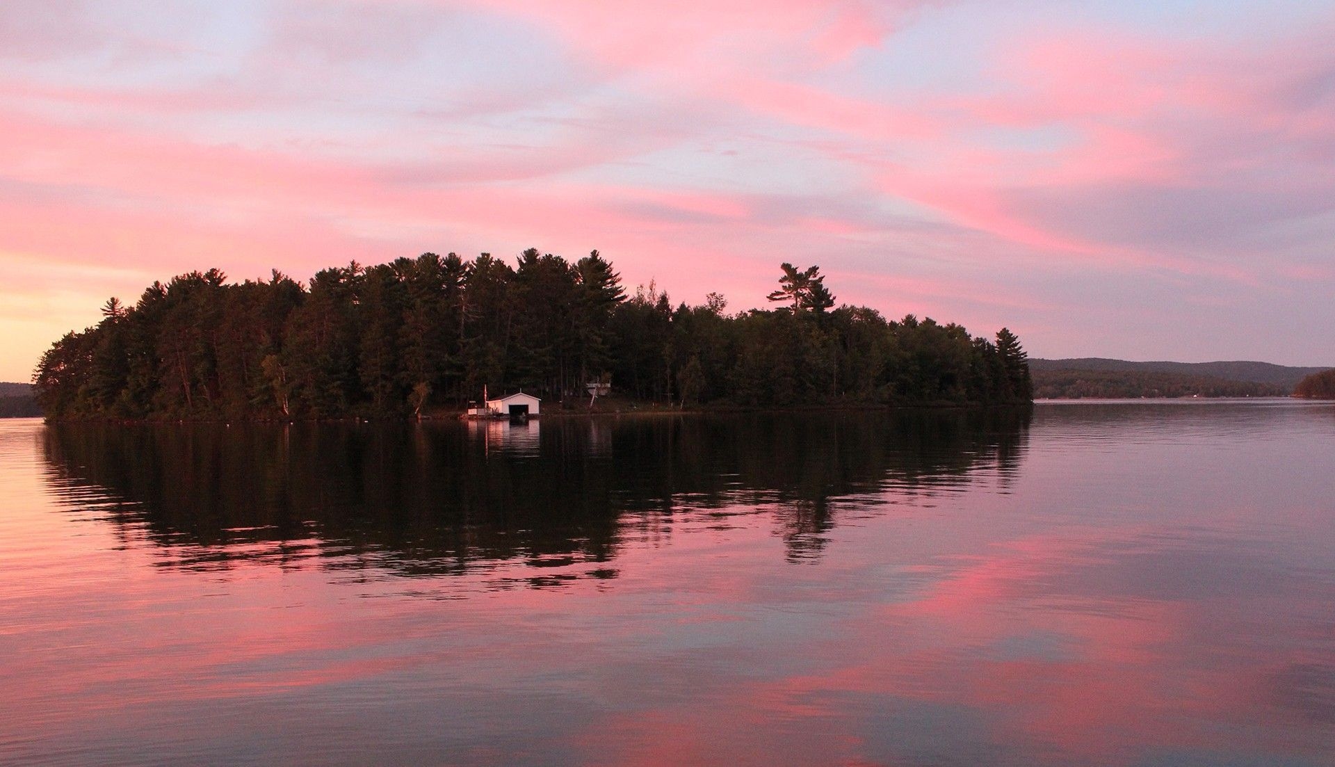 A small cottage on the shore of an island with a pink sunset reflected in the water.