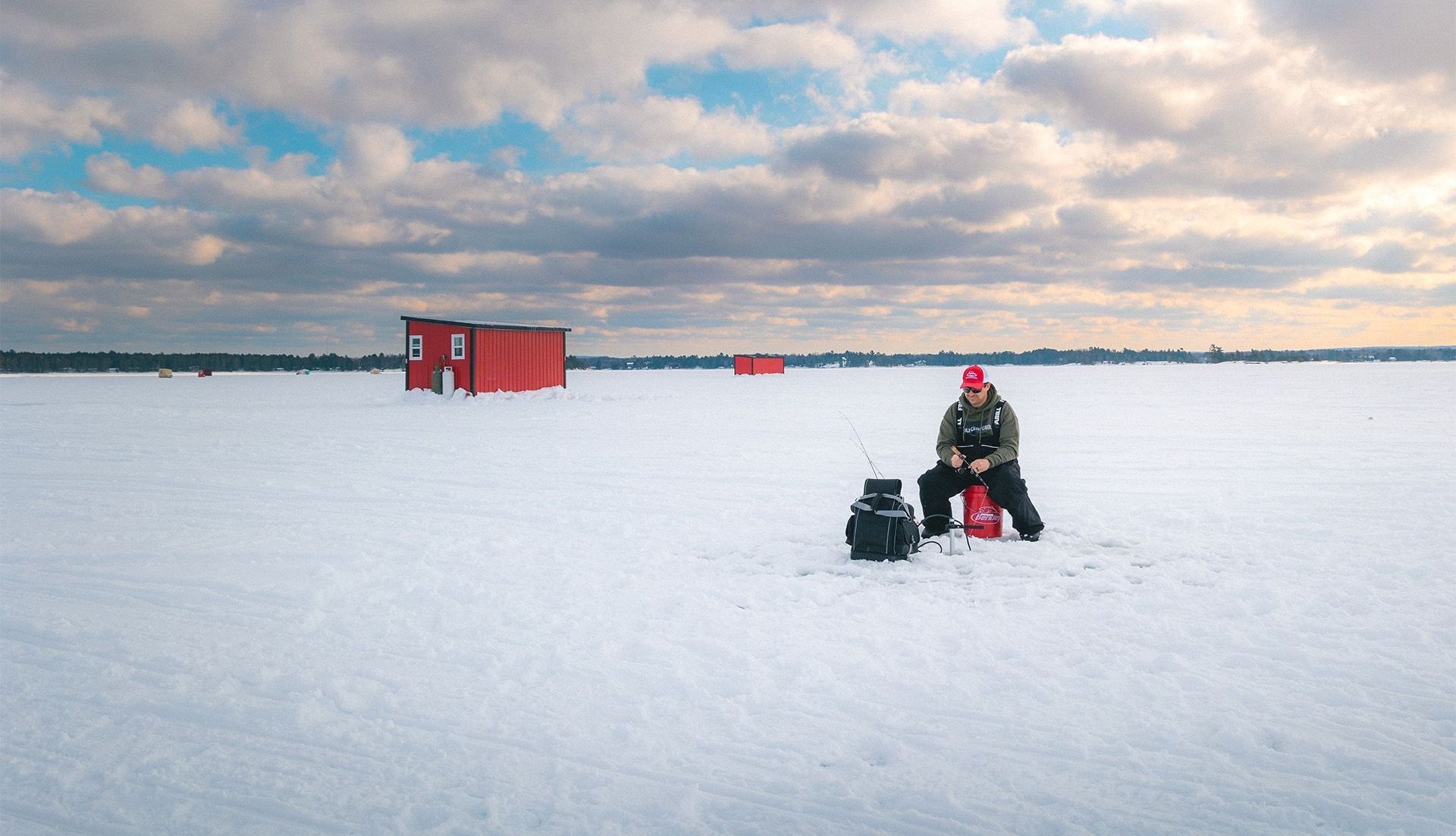 A man ice fishes on a lake in the sunrise near a red fishing hut.