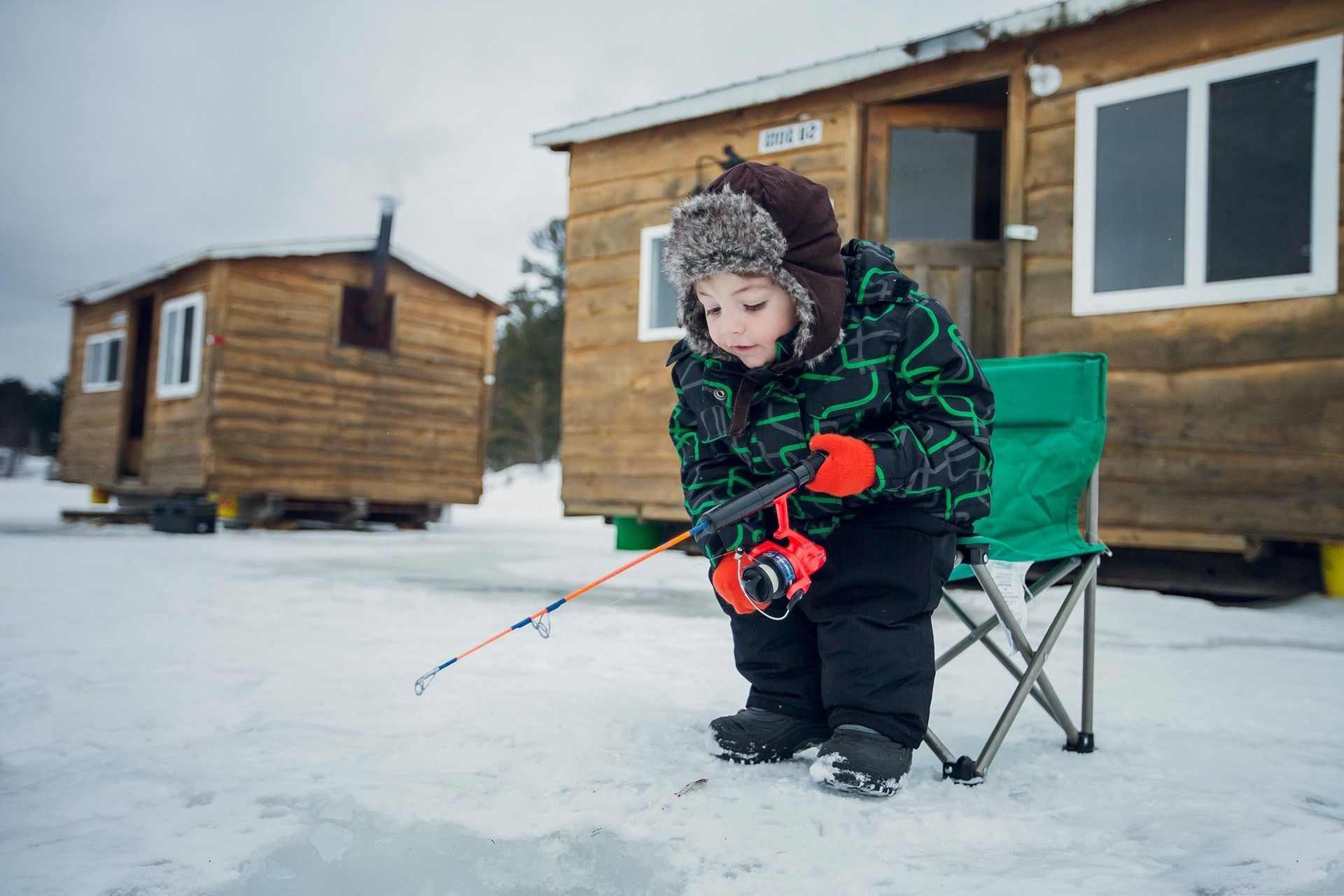 A kid in a bright green and black patterned jacket, and red mits leans over an ice fishing hole with their pole.