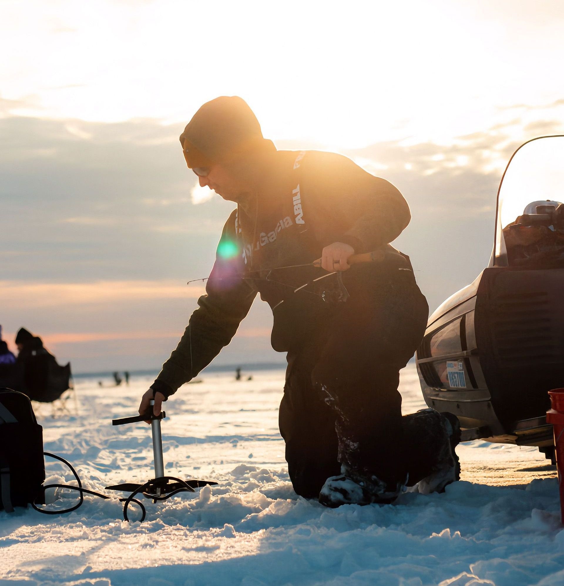 A person in a warm jacket ice fishing at sunset next to their snowmobile.