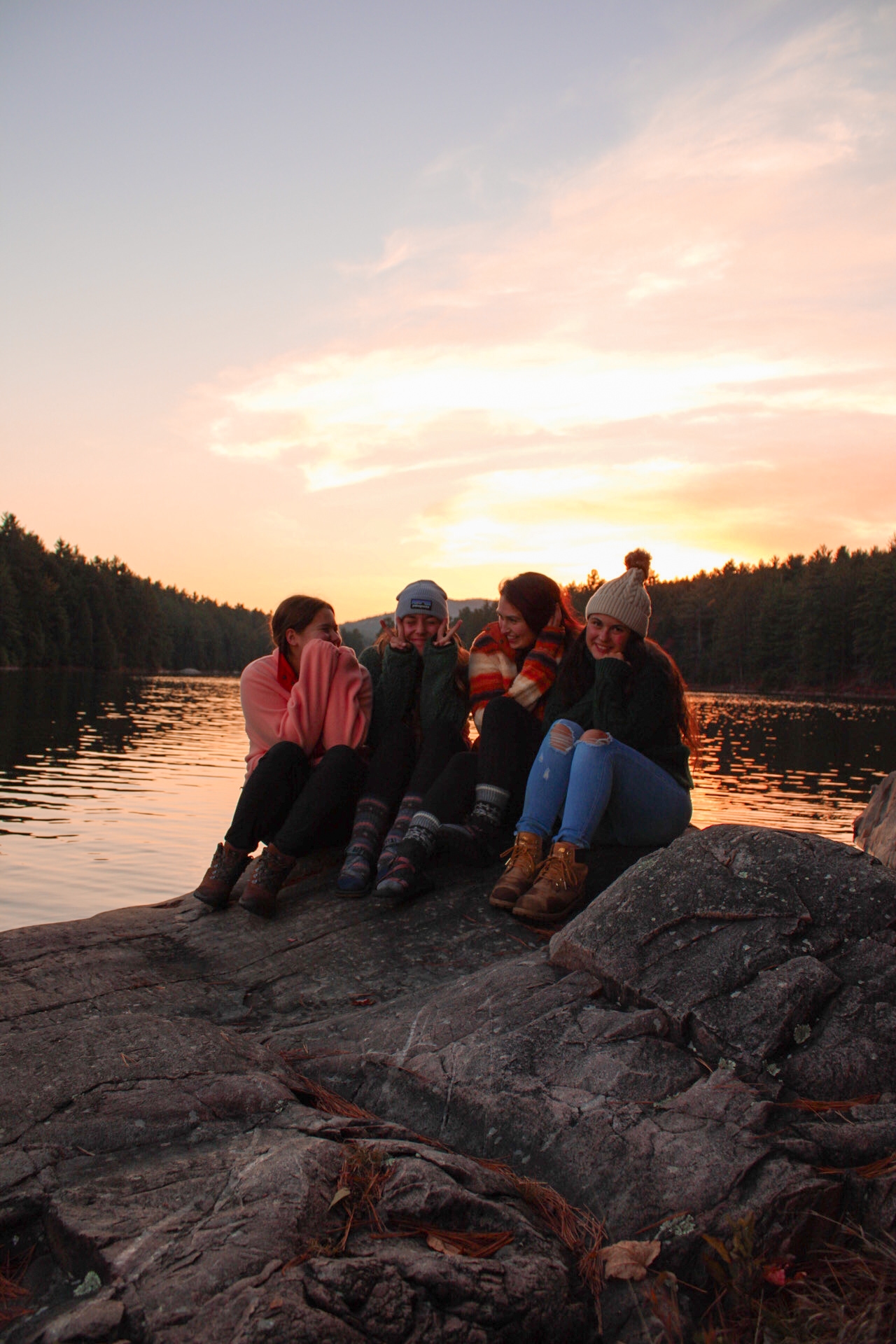 A group of young women on a rock in front of a lake at sunset, smiling and laughing together
