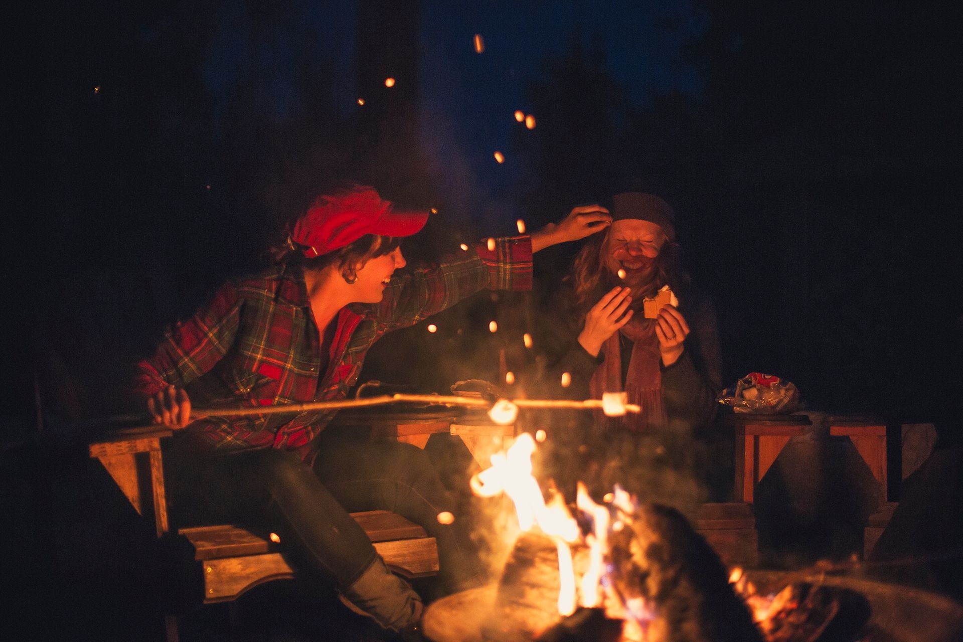 Young people laughing around a bright campfire as one girl takes marshmallow out of another's hair.