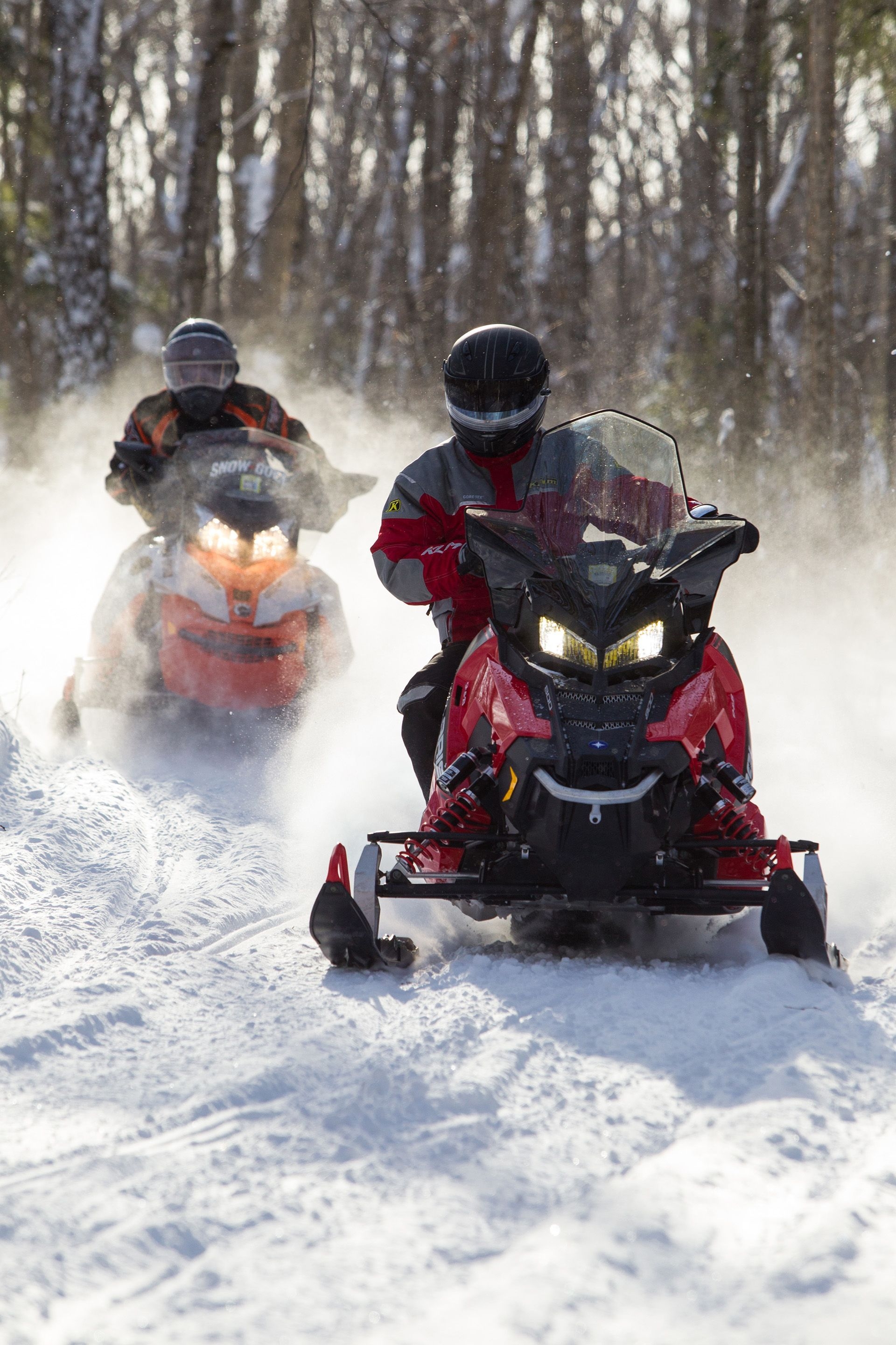 Two people on snowmobiles take a tight turn, kicking up dusty snow powder into the sunlight.