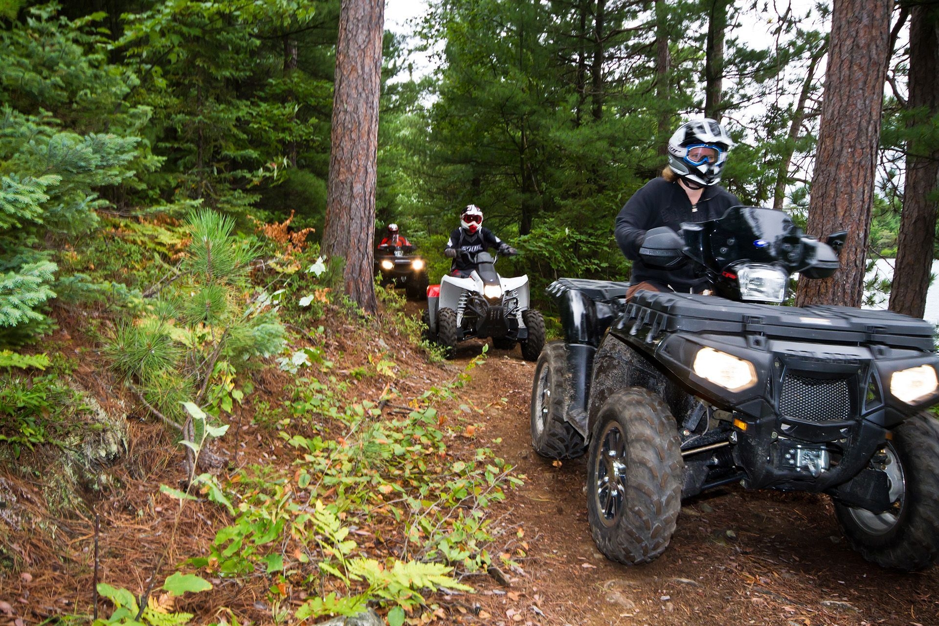 Four people in a line ATVing on a dirt path through the trees.