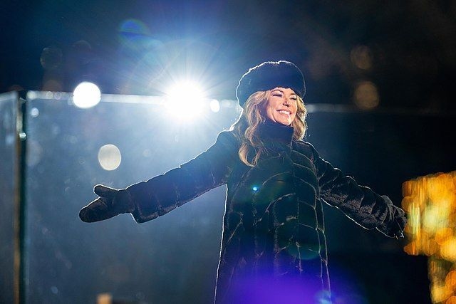 a woman wearing a fur hat and coat smiles and holds her arms out in front of bright lights