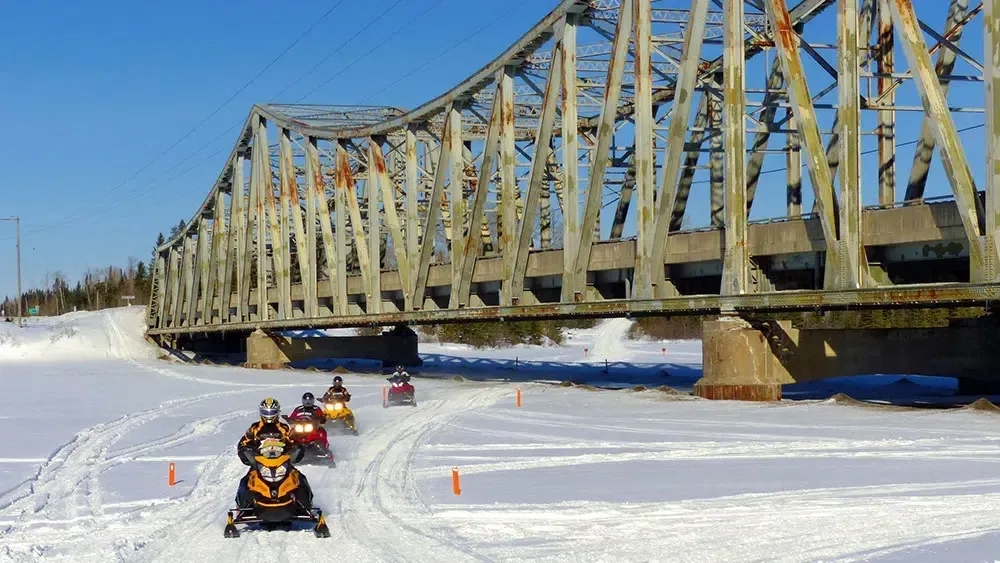 four people on snowmobiles cross a snowy trail underneath a giant steel bridge