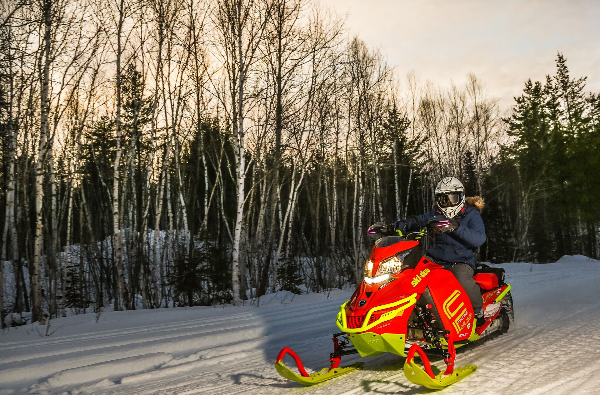 Person riding a snowmobile at dusk on a red and yellow snowmobile.