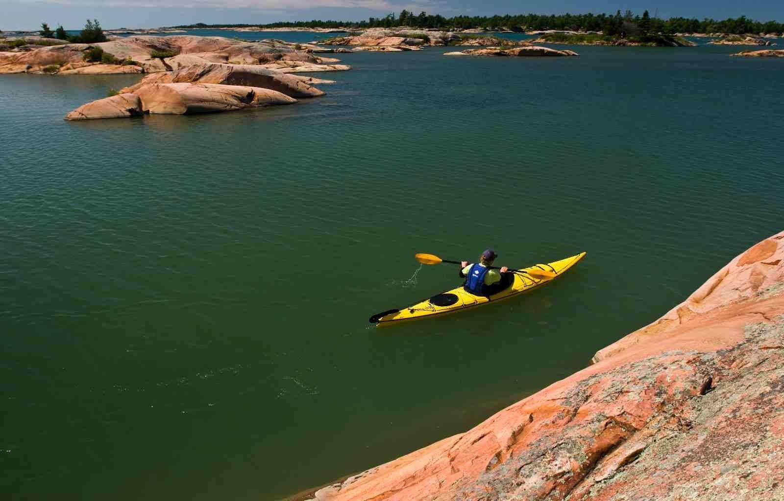 A person in a yellow kayak paddles through teal water near pink rocks in George Lake Killarney.