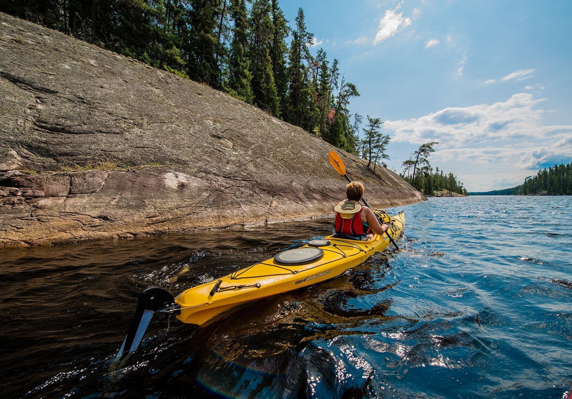 A person in a bright yellowing kayak, paddling next to a sloping rock face at Kaneki Lake near Timmins, Ontario.